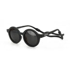 Occhiali da sole zo-oz stile tondo modello Bam nero opaco con fascetta nera per bambini 0-2 anni. Inquadratura laterale in prospettiva con le aste aperte appoggiate su una superficie.
